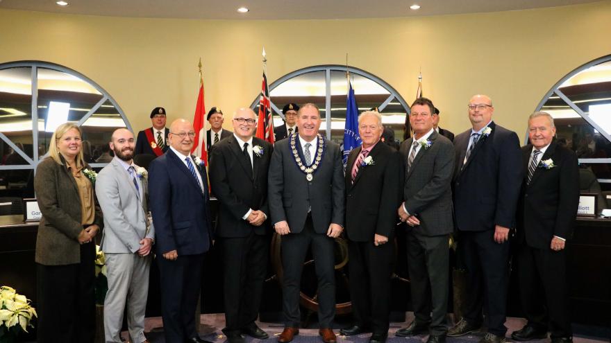 A group photo of individuals in formal attire, standing in front of flags and a council setting.