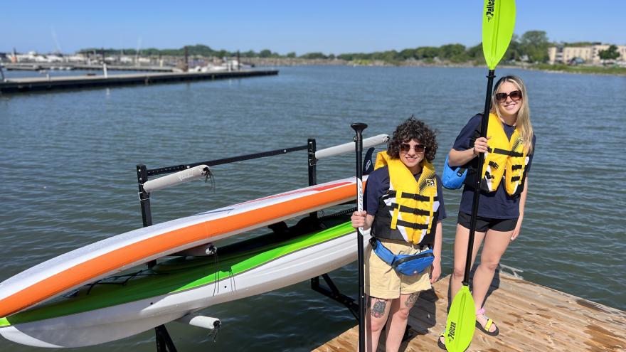 Two individuals wearing life jackets stand on a dock beside a kayak and paddles, with water behind.