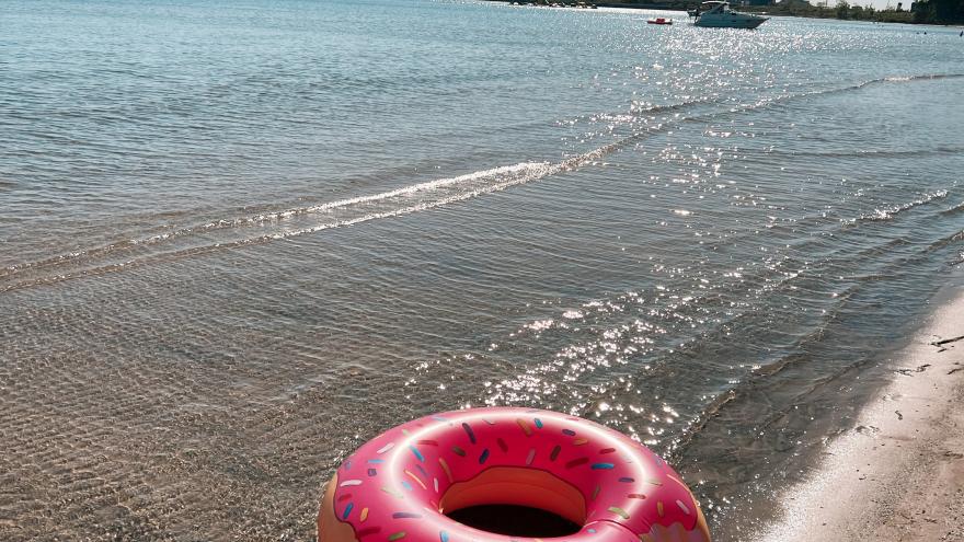 A pink donut-shaped float rests on a sandy beach by the water.