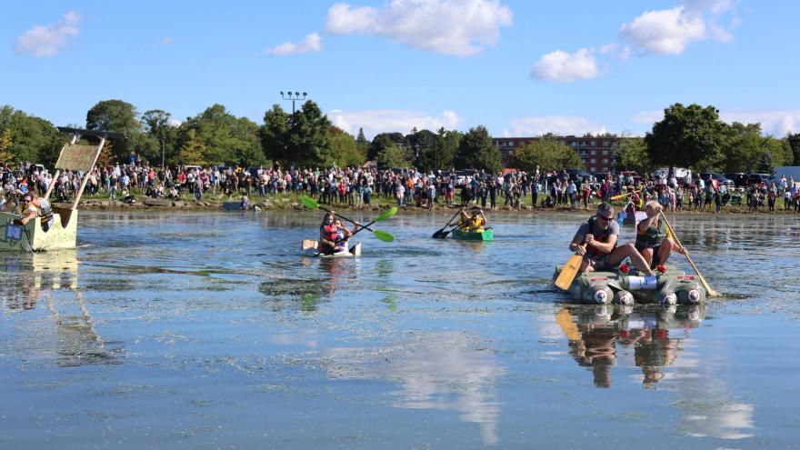 Participants in boats paddle across a calm waterway, surrounded by a large crowd on shore.
