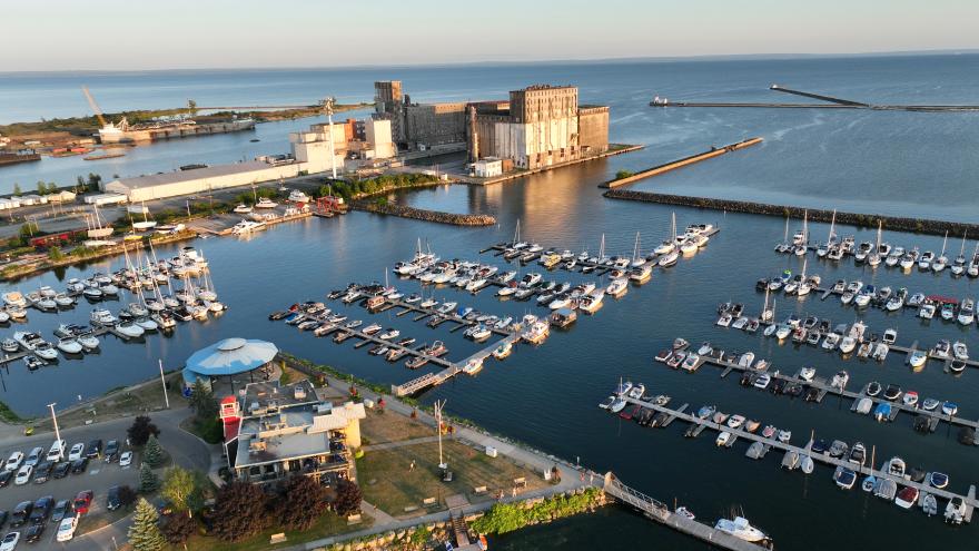 Aerial view of a marina with multiple boats, surrounded by water and a distant industrial area.