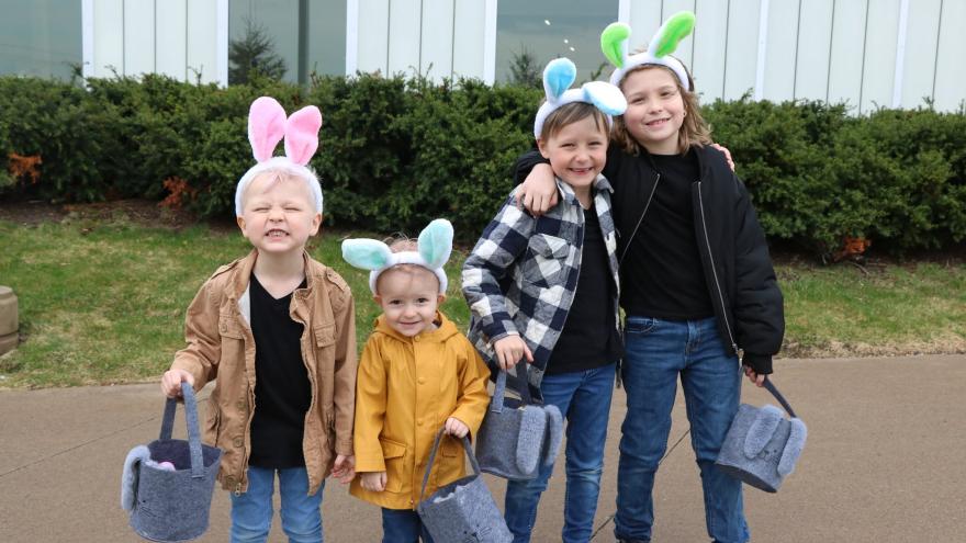 Four children wearing bunny ear headbands pose with baskets outdoors.