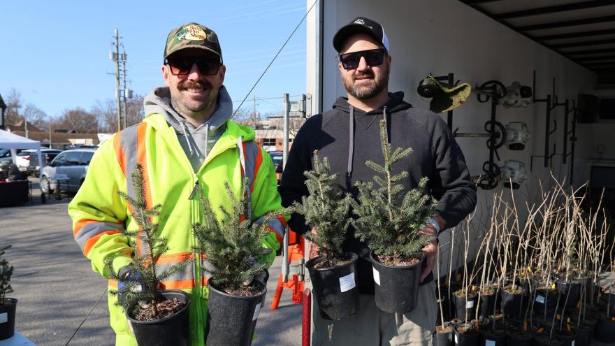 Two individuals hold potted trees, standing outdoors beside a trailer and work equipment.