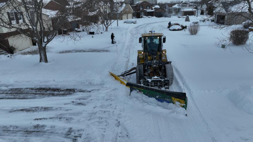 A snowplow clears a snowy residential street with houses in the background.