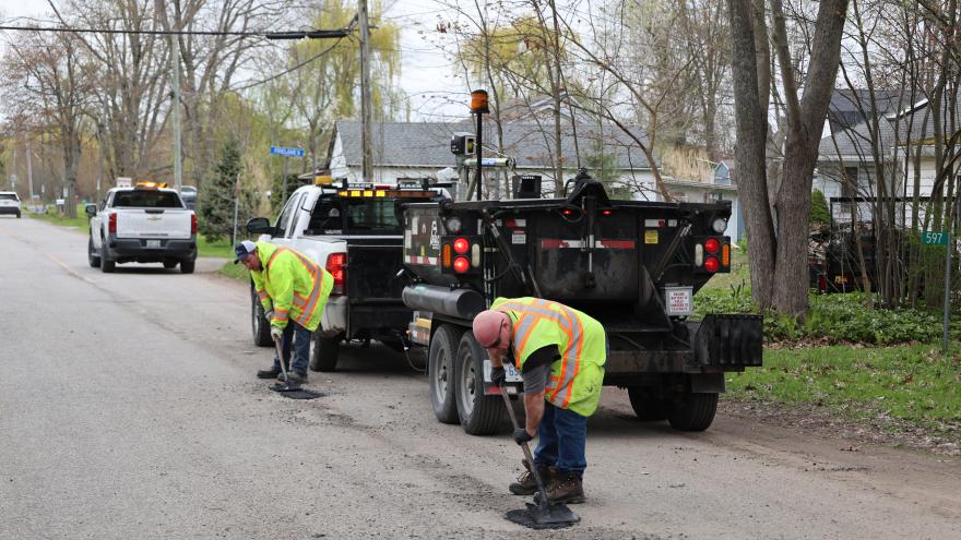 Workers repair a road, using tools beside a parked truck, with trees in the background.