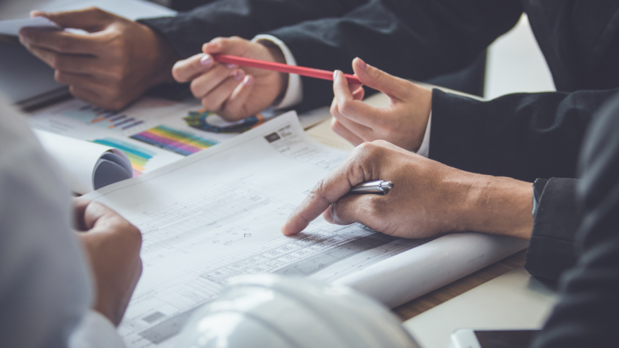 Hands holding pens and pointing at a document with graphs on a table. A construction helmet is visible.