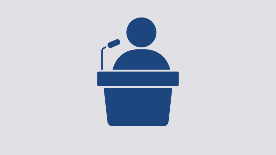 Silhouette of a person at a podium with a microphone on a light background.