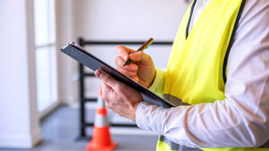 A person in a high-visibility vest takes notes on a clipboard in a well-lit indoor setting.