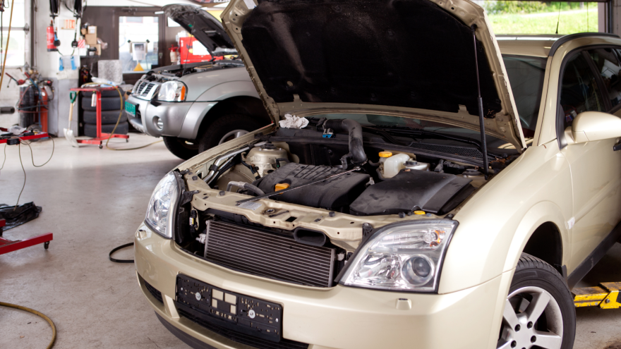 A silver car with its hood open is in a garage, with a second vehicle in the background.