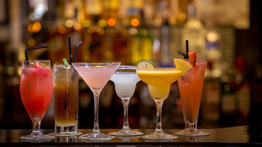 A row of colorful cocktails on a bar with a blurred background of bottles.