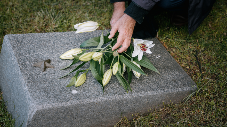 A hand places a bouquet of white lilies on a grey gravestone in a grassy area.
