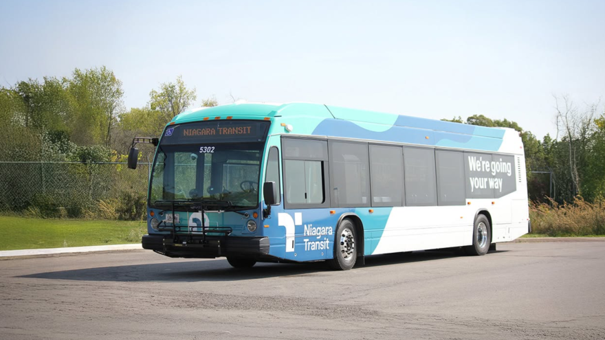 City bus stopped on a paved surface, surrounded by green trees and shrubs.