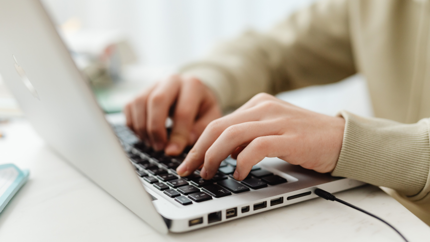 Hands typing on a laptop keyboard, sitting on a light-coloured table.