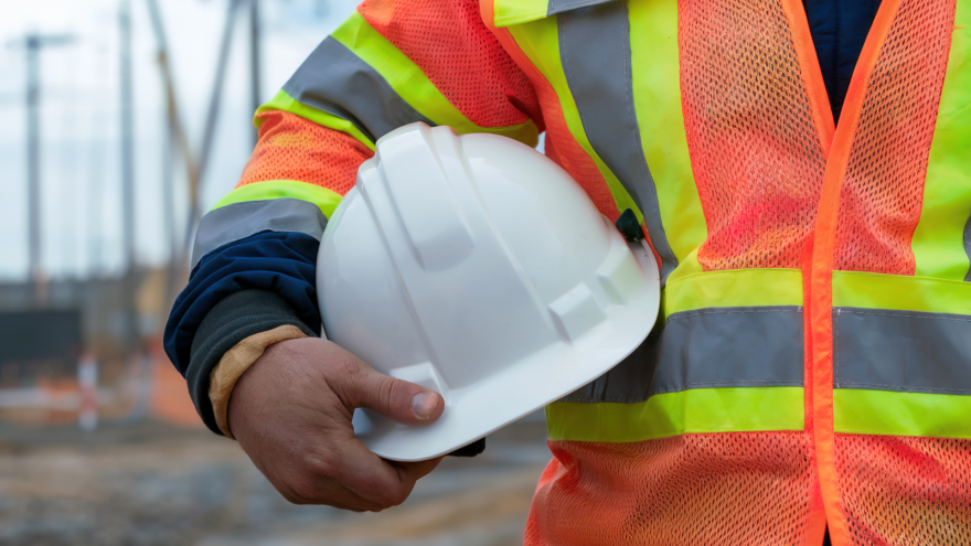 A person in a safety vest holding a white hard hat at a construction site.