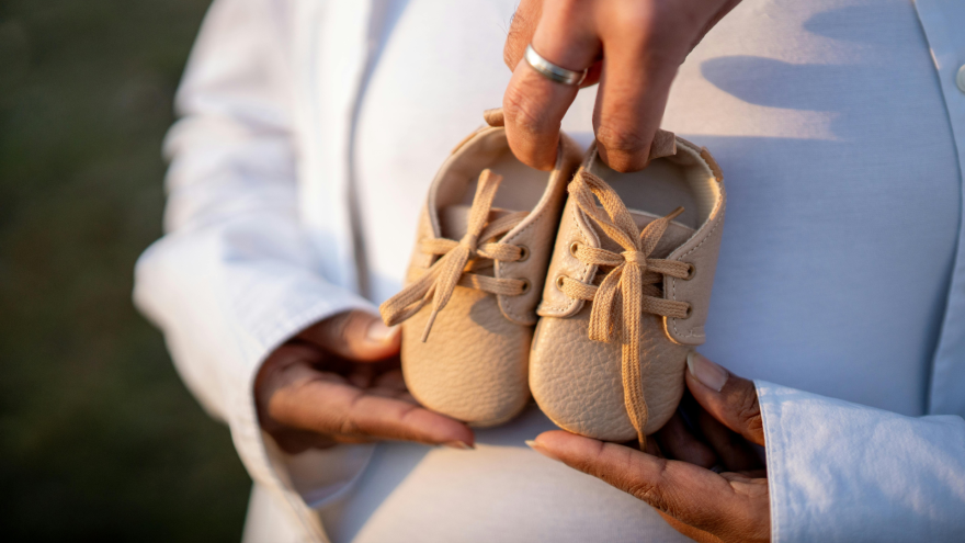 Hands holding tiny beige baby shoes in front of a pregnant person's belly.