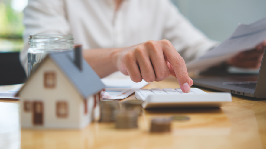 A person using a calculator with documents and a model house on a desk.