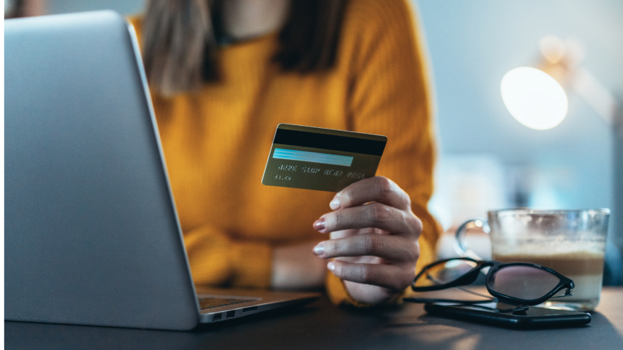 A hand holding a credit card in front of a laptop, with a coffee cup and glasses nearby.