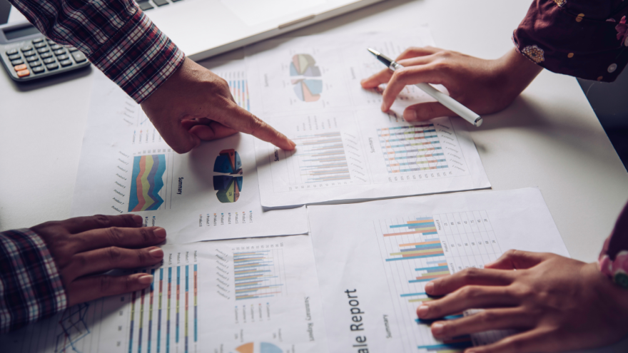 Hands pointing at printed graphs and charts on a table, with a calculator nearby.