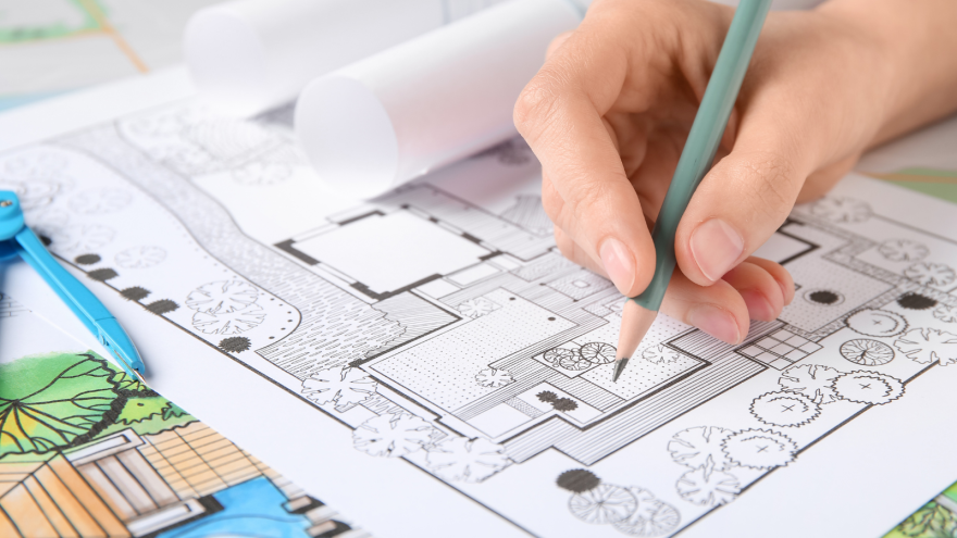 A hand holding a pencil sketches landscaping designs on a blueprint surrounded by rolled plans.