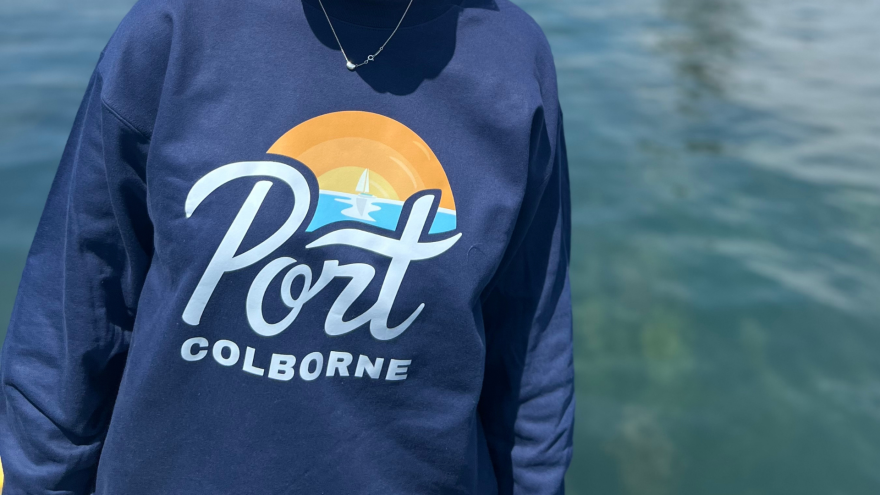 A person wearing a navy sweatshirt with "Port Colborne" and a sunset design, near water.
