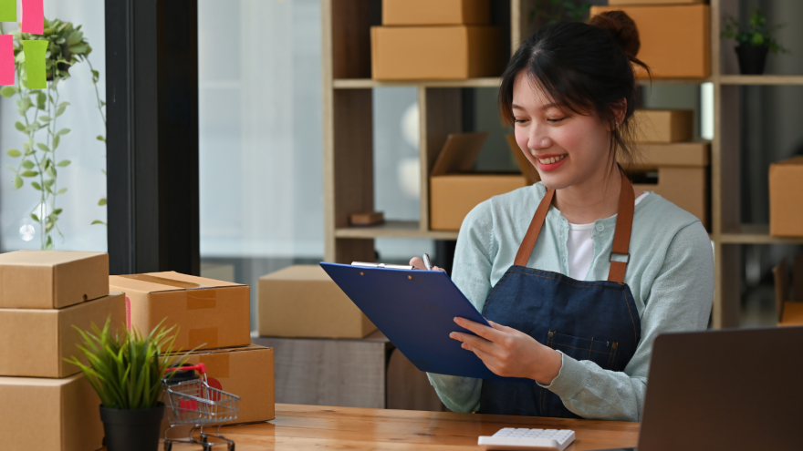 A person with a clipboard smiles while sitting at a desk surrounded by boxes.