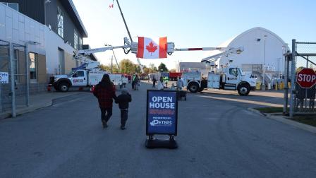 Two people walk towards a sign for an open house, with Canadian flags in the background.
