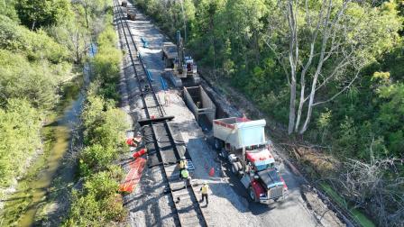 Construction site with machinery and workers beside a road, surrounded by trees and water.