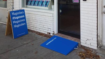 Blue accessibility ramp leading to a shop entrance with a sign promoting magazines.