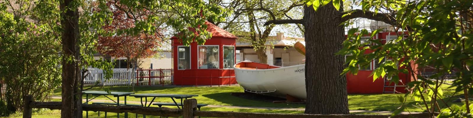 A sunny park scene featuring red structures and a white boat among green trees and picnic tables.