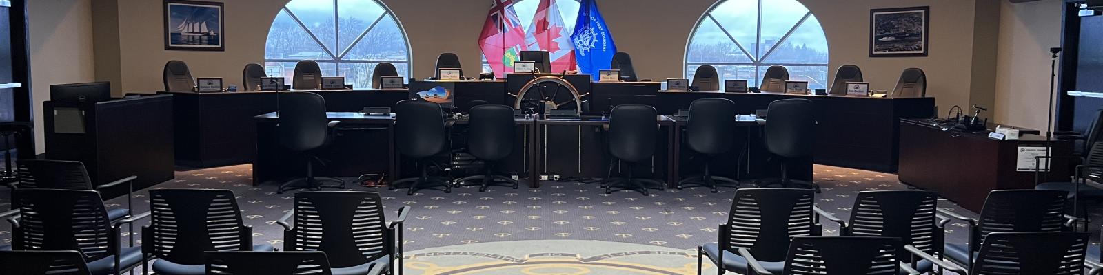 Council chamber with empty chairs, flags, and a circular decorative floor design.