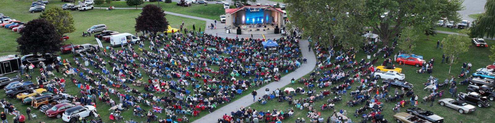 Aerial view of a large outdoor concert with a crowd sitting on grass and classic cars parked nearby.