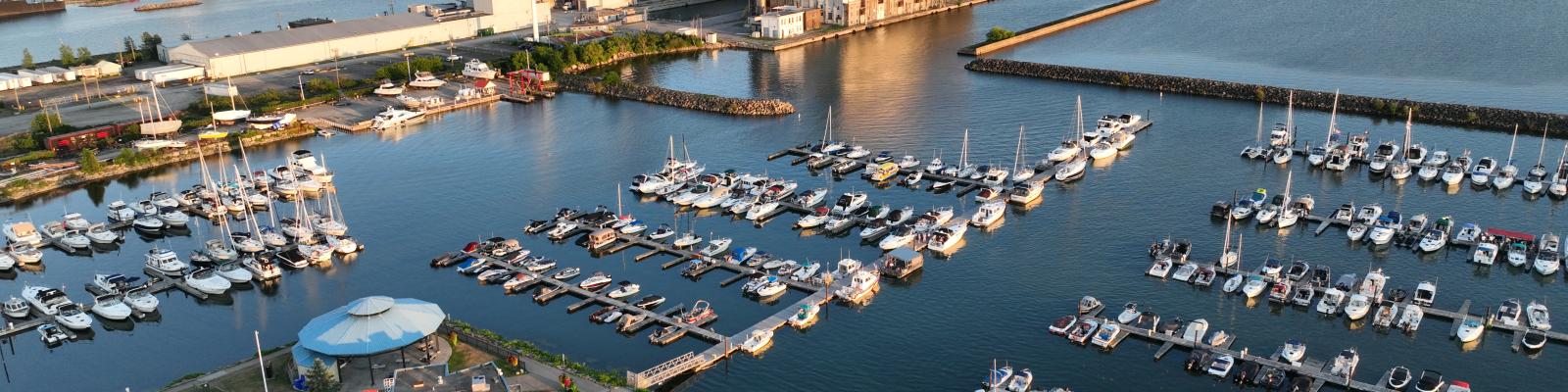 Aerial view of a marina with multiple boats, surrounded by water and a distant industrial area.