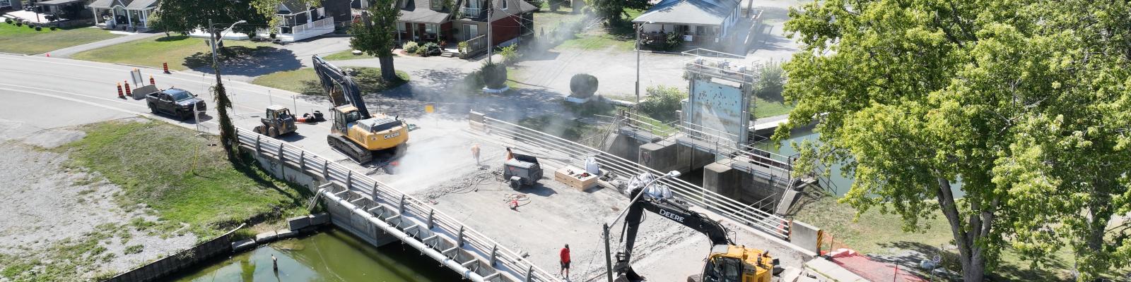 Construction equipment and workers on a bridge; smoke and vehicles visible, surrounded by greenery.