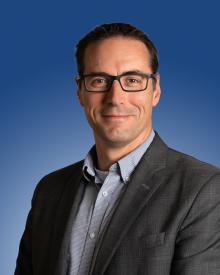 Professional portrait against a blue background, featuring a smiling individual in a suit and glasses.