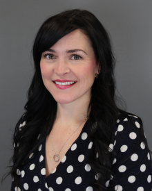 Person with long dark hair wearing a polka dot blouse, smiling against a grey background.
