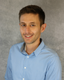 Smiling person wearing a blue checkered shirt against a grey background.