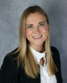 Smiling individual with long, straight hair, wearing a black blazer and white shirt against a grey background.