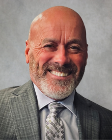 A smiling person in a grey suit with a checkered tie against a neutral background.