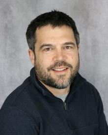 Smiling individual with a beard, wearing a dark sweater, against a grey background.