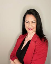 Smiling person with long dark hair, wearing a red blazer and black top, standing against a neutral background.