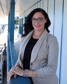 Person with glasses smiling while leaning on a blue railing, with a seaside background.