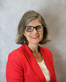 A smiling individual in a red blazer poses against a grey background.