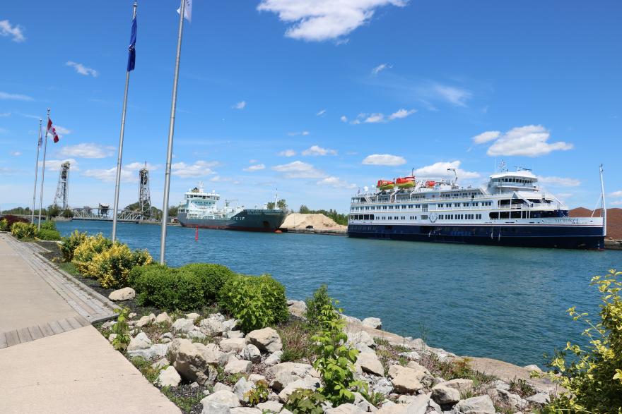 Two large ships docked in a sunny harbour, with flags and greenery nearby.