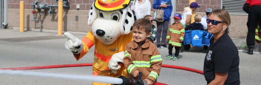 A child in a fire suit smiles, holding a hose with a mascot beside them at a community event.