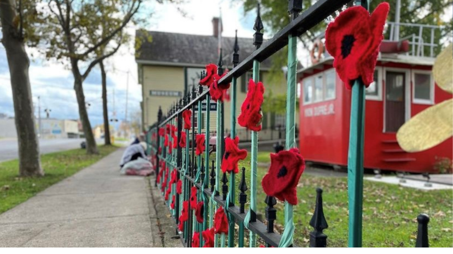 Knitted red poppies adorn a green fence beside a building and a red structure.