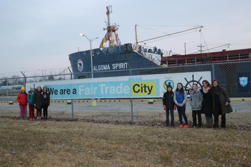 Group stands near a sign reading "We are a Fair Trade City" with a ship in the background.