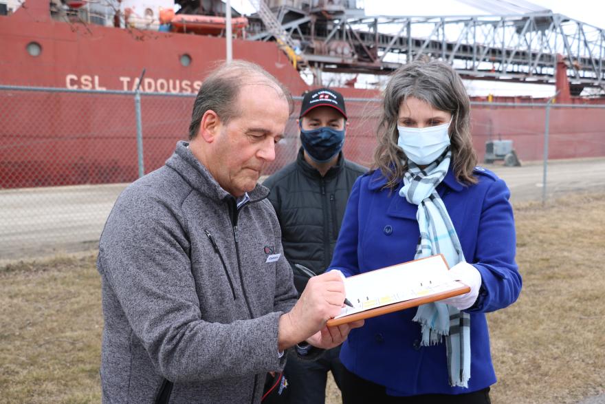 Two individuals stand outdoors, one signing a document while another observes, with a ship in the background.