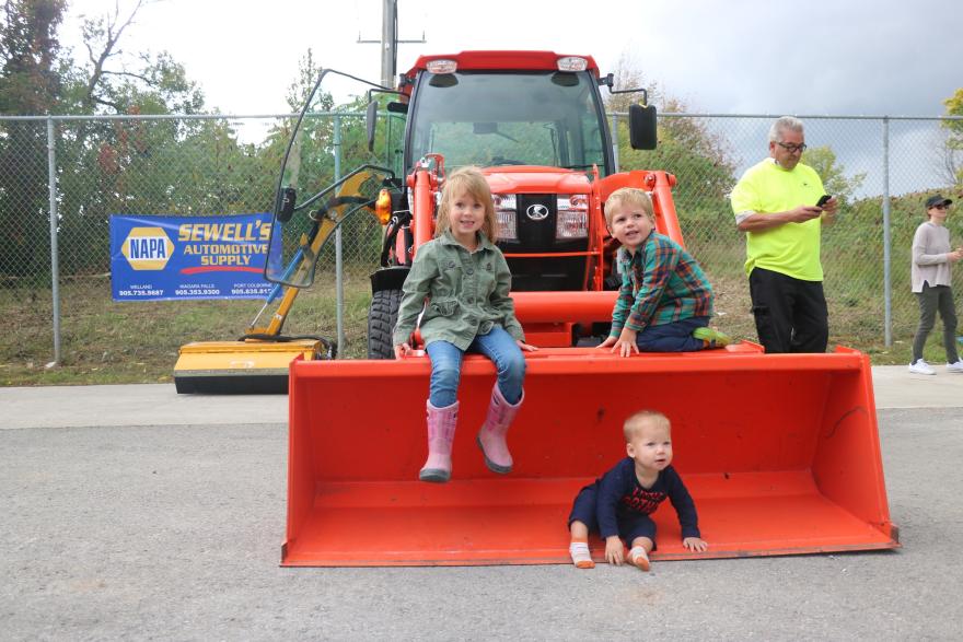 Two children sit on an oversized orange equipment bucket, with a baby crawling nearby. A tractor is in the background.