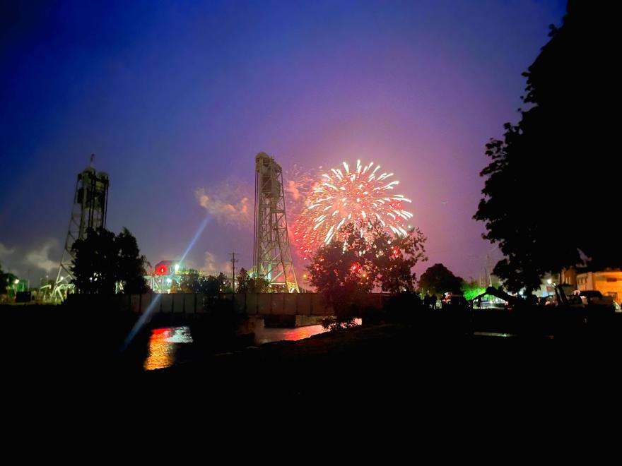 Fireworks illuminate the night sky, with structures and trees silhouetted in the foreground.