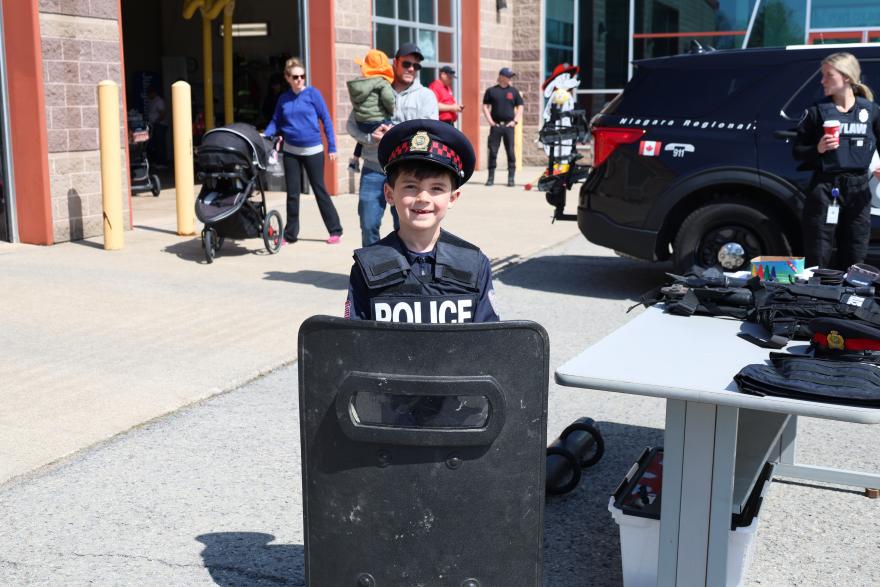 A child in a police uniform smiles behind a shield at a community event, with bystanders in the background.